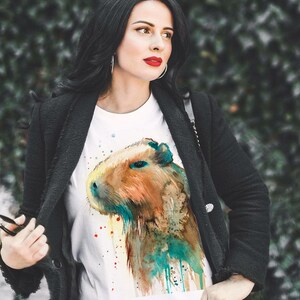 May include: White t-shirt with a watercolor illustration of a capybara. The capybara is brown and has a blue and green watercolor background.