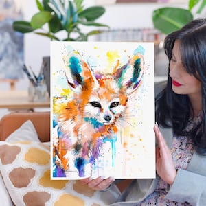 Puede incluir: Pintura acuarela de un zorro fennec con pelaje naranja, orejas grandes y ojos negros. La pintura tiene un fondo colorido con salpicaduras de amarillo, azul y morado.
