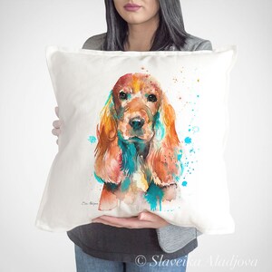 Può includere: Un cuscino bianco con un dipinto ad acquerello di un cocker spaniel. Il cane è marrone e bianco con accenti blu e verdi. Il cuscino è decorato con schizzi di acquerello blu e verdi.