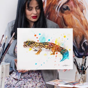 May include: A watercolor painting of a gecko with a colorful tail and spots of color around it. The painting is on white paper and is being held by a person.