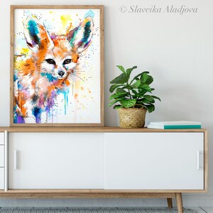 Puede incluir: Una pintura de acuarela de un zorro fennec con orejas grandes, sobre un fondo blanco con salpicaduras de pintura amarilla, azul y morada.