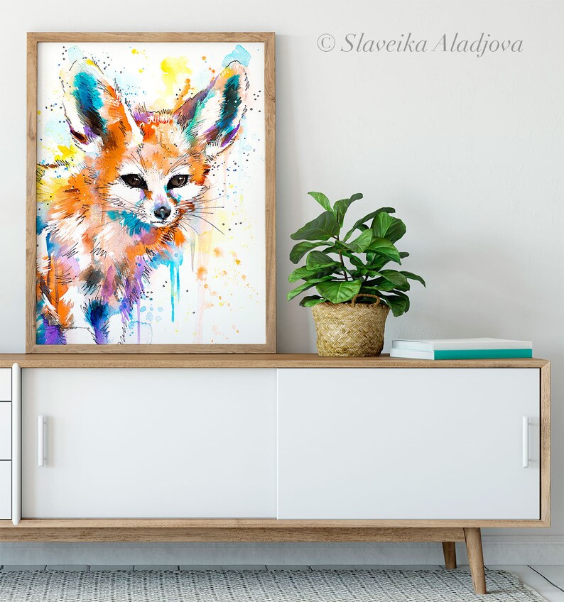 Puede incluir: Una pintura de acuarela de un zorro fennec con orejas grandes, sobre un fondo blanco con salpicaduras de pintura amarilla, azul y morada.