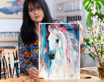 May include: A watercolor painting of a white horse with a blue, pink, and red background. The horse's head is turned to the side, and its mane is flowing in the wind. The painting is framed and ready to hang.