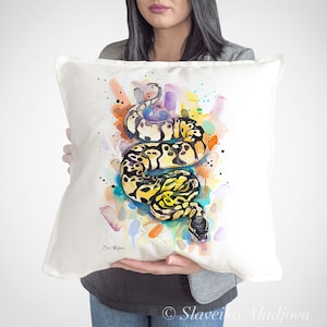 Op de afbeelding: Een wit kussen met een aquarel schilderij van een gele en zwarte slang op een kleurrijke achtergrond. De slang is opgerold in een spiraalvorm. De achtergrond is een mix van oranje, geel, blauw, paars en groen.