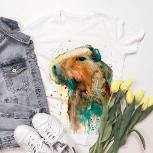 May include: White t-shirt with a watercolor illustration of a capybara. The capybara is brown and orange with blue and green watercolor accents. The background is white with splashes of red, blue, and yellow watercolor.