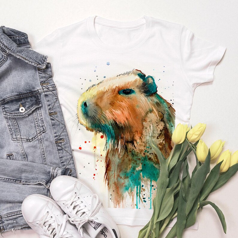 May include: White t-shirt with a watercolor illustration of a capybara. The capybara is brown and orange with blue and green watercolor accents. The background is white with splashes of red, blue, and yellow watercolor.