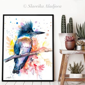May include: Watercolor painting of a blue and brown kingfisher perched on a branch with colorful flowers. The bird has a crest of feathers and a long beak. The background is white with scattered blue and pink dots.
