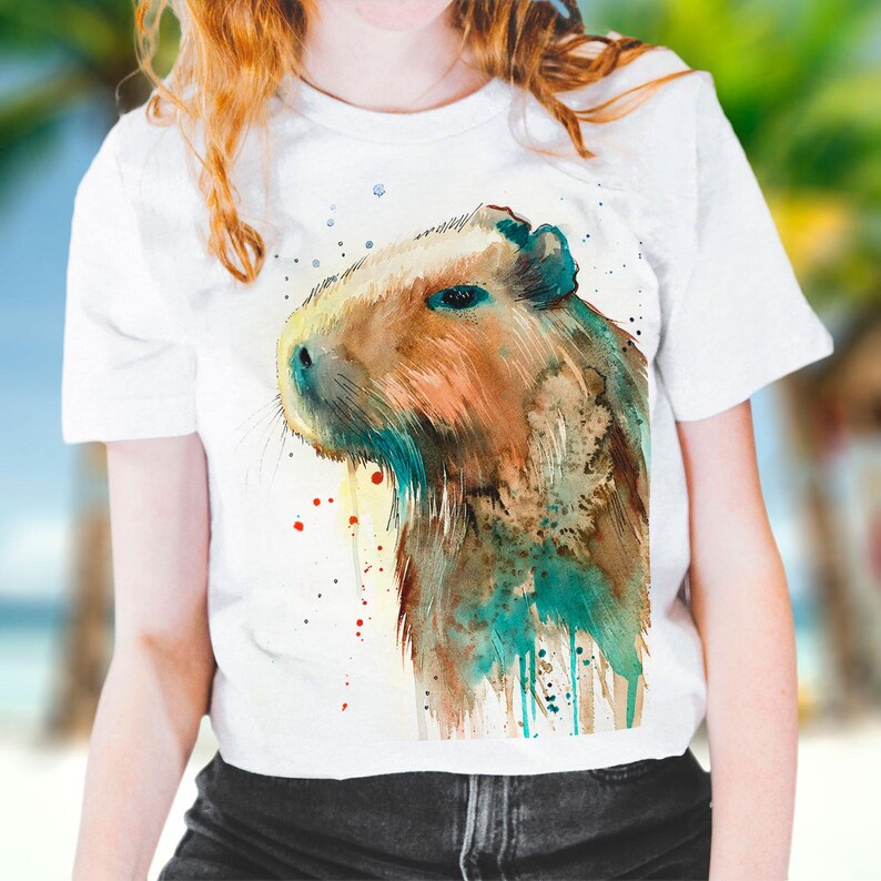 May include: White t-shirt with a watercolor illustration of a capybara. The capybara is brown and has a blue and green watercolor effect around it.