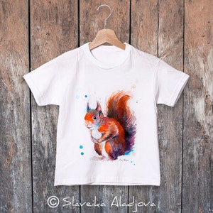 May include: White t-shirt featuring a watercolour painting of a red squirrel. The squirrel is standing on its hind legs with its tail curled around its body. The background is a light blue with white dots.