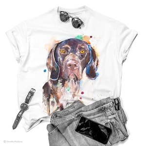 May include: A white t-shirt with a watercolor illustration of a brown and white dog with yellow eyes. The dog is surrounded by colorful watercolor splatters. The t-shirt is laid out on a white surface with a pair of sunglasses, a watch, and a pair of jeans.