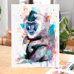 May include: Watercolor painting of a mandrill monkey with blue, purple, and orange hues. The monkey is sitting with its arms crossed and looking directly at the viewer.