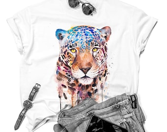 May include: A white t-shirt with a colorful watercolor illustration of a jaguar's face. The jaguar has blue, green, yellow, orange, and brown markings. The shirt is laid flat on a white background with a pair of sunglasses, a watch, and a pair of jeans.