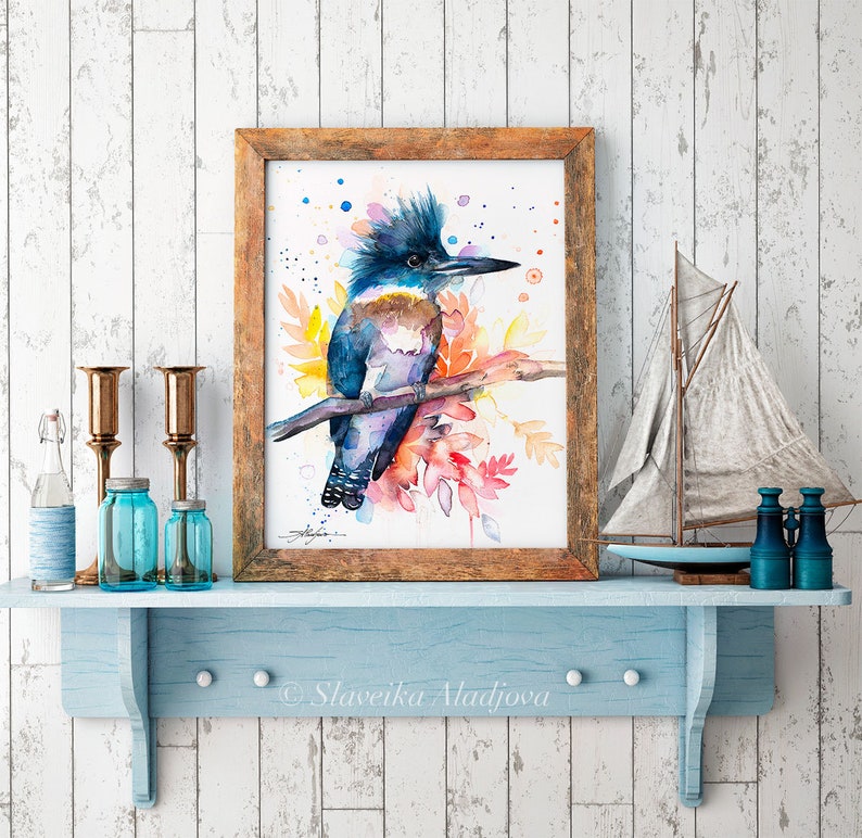 May include: Watercolor painting of a blue and purple belted kingfisher perched on a branch with colorful watercolor leaves. The bird has a crested head and a long, pointed beak. The painting is framed in a rustic wooden frame.