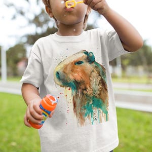 May include: A white t-shirt with a watercolor illustration of a capybara. The capybara is brown and has a green and blue watercolor effect around it.