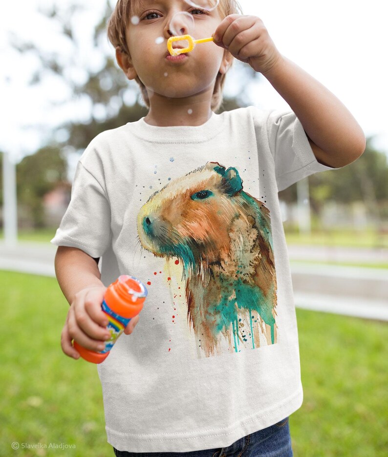 May include: A white t-shirt with a watercolor illustration of a capybara. The capybara is brown and has a green and blue watercolor effect around it.