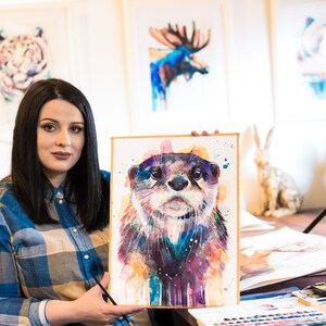 May include: A woman holds up a watercolor painting of an otter. The otter is painted in shades of blue, purple, and yellow. The painting is framed in a simple wooden frame.