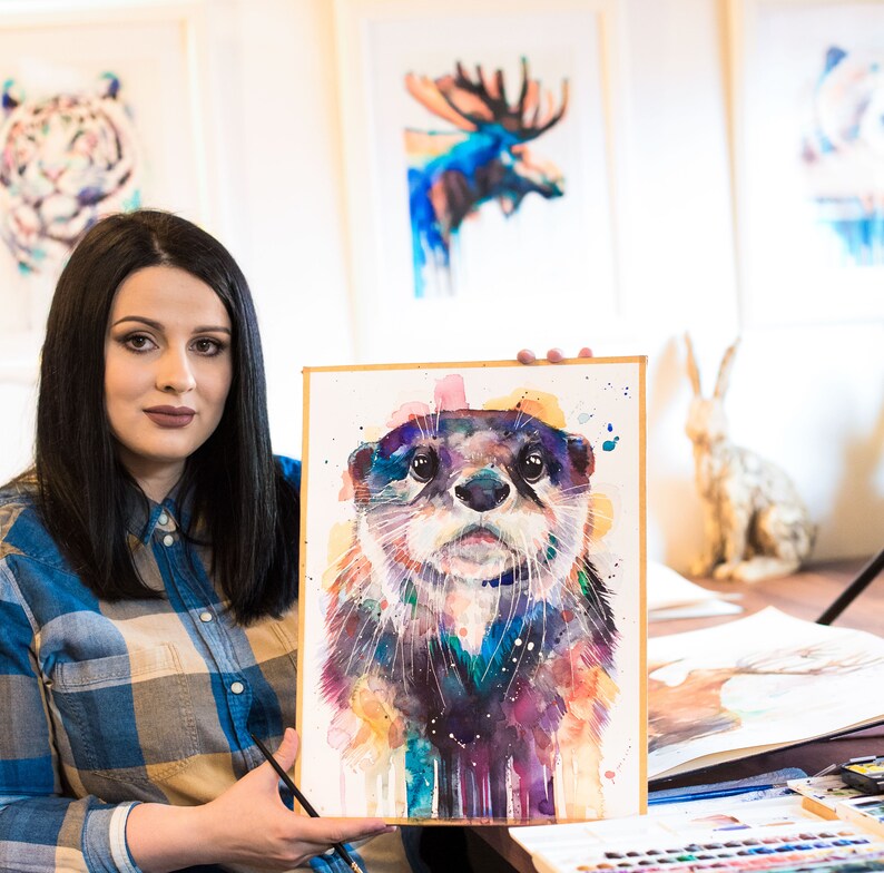 May include: A woman holds up a watercolor painting of an otter. The otter is painted in shades of blue, purple, and yellow. The painting is framed in a simple wooden frame.