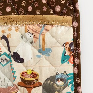 May include: A quilted pot holder featuring a cat-themed fabric. The design includes various cats in hats and scenes, with a brown paw print border and a cork accent. The pot holder is quilted with white thread.