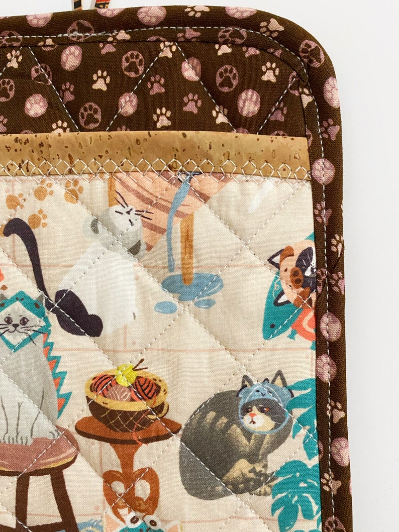 May include: A quilted pot holder featuring a cat-themed fabric. The design includes various cats in hats and scenes, with a brown paw print border and a cork accent. The pot holder is quilted with white thread.