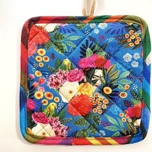 Puede incluir: Un posavasos acolchado azul con un estampado floral colorido que presenta el rostro de una mujer, pájaros y flores. El posavasos tiene un asa de cuero marrón.