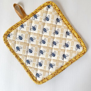 May include: A quilted, square-shaped pot holder with a bee and leaf pattern. The main fabric is off-white with a repeating pattern of blue bees and gold leaves. The border is a honeycomb pattern in gold, and it has a brown loop for hanging.