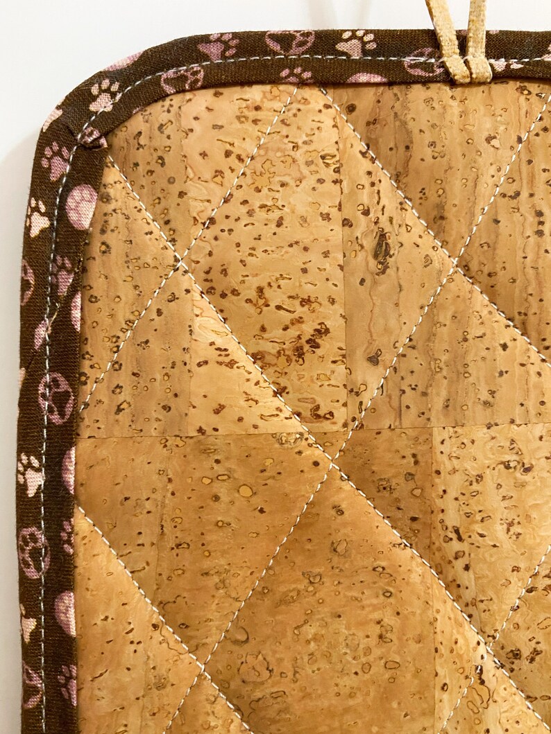 May include: A quilted cork hot pad with a brown paw print border and a beige loop for hanging. The pad features a diamond pattern created by white stitching. The cork has a natural, speckled appearance.