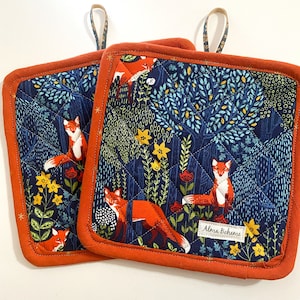 May include: Two square quilted pot holders with a navy blue background featuring orange foxes, yellow flowers, and green foliage. The pot holders have an orange border and a hanging loop. The text "SAMPLE WITHOUT A POCKET" is at the top.