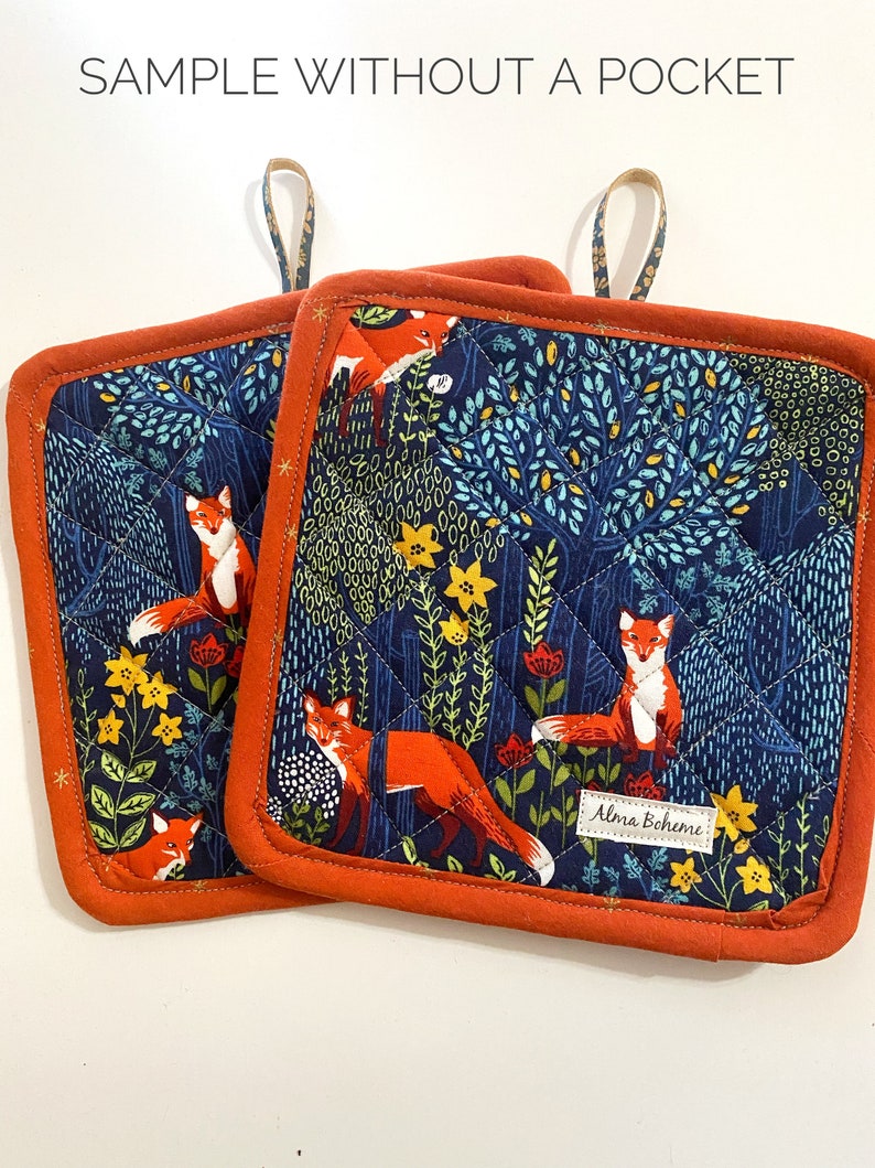 May include: Two square quilted pot holders with a navy blue background featuring orange foxes, yellow flowers, and green foliage. The pot holders have an orange border and a hanging loop. The text "SAMPLE WITHOUT A POCKET" is at the top.