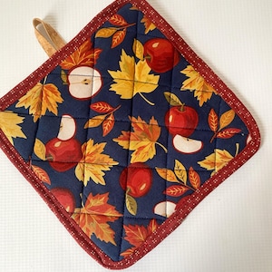 May include: A square quilted potholder featuring a navy blue background with a repeating pattern of red apples, apple slices, and yellow and orange autumn maple leaves. The edges are finished with a dark red fabric trim and a small hanging loop.