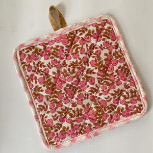 May include: A square quilted potholder featuring a repeating pattern of pink apples and brown leaves on a white background. The edges are finished with a pink and white gingham border, and a small tan cork loop is attached to the top corner.