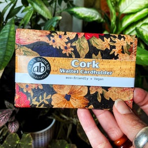 May include: A cork wallet cardholder with a floral pattern in red, orange, and yellow on a black background. The wallet has a white band with the words "Cork Wallet Cardholder" and "eco-friendly * Vegan". The wallet is held in a hand.