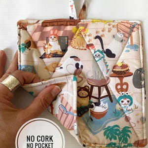 May include: A quilted beige and cream pot holder with a loop for hanging. The fabric features a repeating pattern of cartoon cats in various poses and costumes. The pot holder is square with a diamond-stitched pattern.