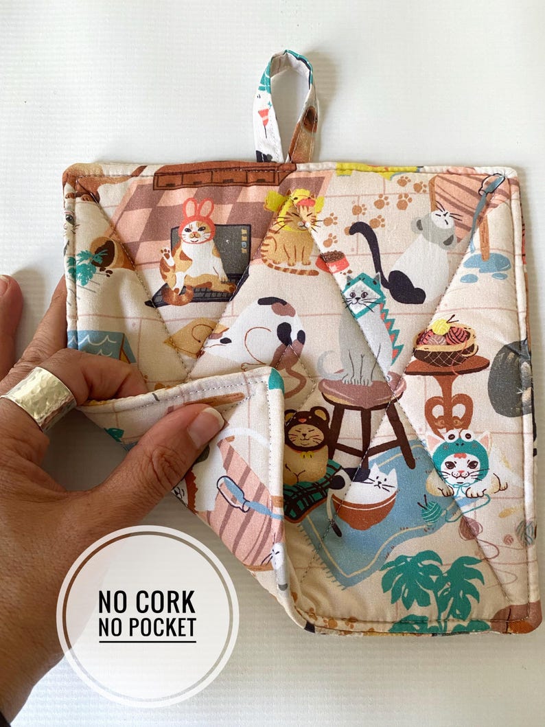 May include: A quilted beige and cream pot holder with a loop for hanging. The fabric features a repeating pattern of cartoon cats in various poses and costumes. The pot holder is square with a diamond-stitched pattern.