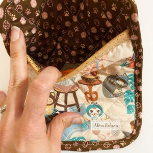 May include: A quilted oven mitt with a brown paw print pattern on the inside and a cat-themed fabric on the front. The mitt has a brown border and the label "Alma Boheme".