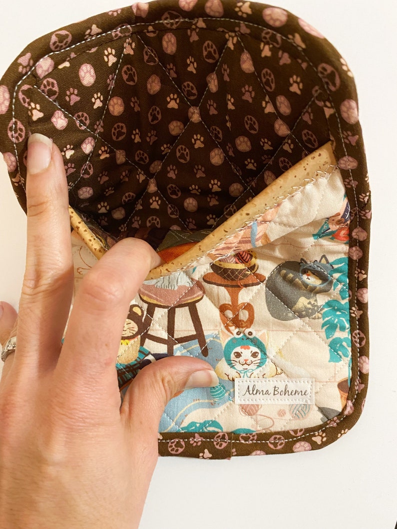 May include: A quilted oven mitt with a brown paw print pattern on the inside and a cat-themed fabric on the front. The mitt has a brown border and the label "Alma Boheme".