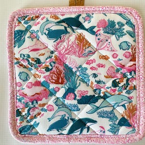 Puede incluir: Salvamanteles cuadrado acolchado con un estampado de vida marina. El diseño presenta delfines, tortugas marinas, peces y corales en tonos turquesa, rosa y naranja sobre un fondo blanco. Los bordes están ribeteados con tela floral rosa y un lazo de corcho.