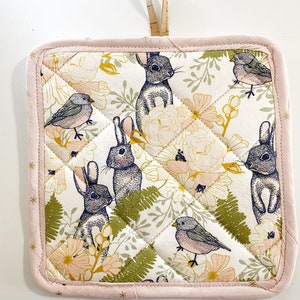 May include: A pink quilted hot pad with a floral pattern featuring bunnies, birds, and leaves. The pattern is in shades of blue, green, yellow, and pink.