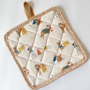 Puede incluir: Salvamanteles cuadrado acolchado con fondo crema y un estampado repetido de perros con suéteres y llevando bolsas. El salvamanteles tiene un borde floral marrón y un lazo para colgar.