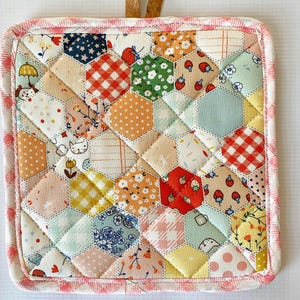 May include: A quilted square hot pad with a patchwork design. The pad features a variety of hexagonal fabric pieces in colors like red, orange, green, and blue, with patterns including strawberries, polka dots, and floral prints. A pink gingham border and a cork loop complete the design.