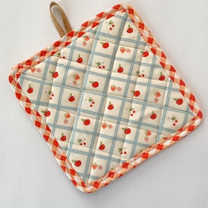 May include: Square quilted pot holder with a fruit pattern. The center features a cream-colored fabric with a blue grid pattern and red apples, cherries, and strawberries. The border is a red and orange gingham pattern, and it has a brown loop for hanging.