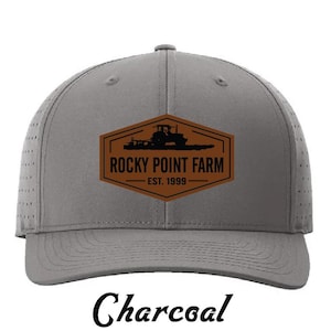 May include: Charcoal-colored baseball cap featuring a brown and black Rocky Point Farm logo with a tractor silhouette. The logo includes the text "Rocky Point Farm" and "EST. 1999". The word "Charcoal" is written in a cursive font at the bottom of the cap.