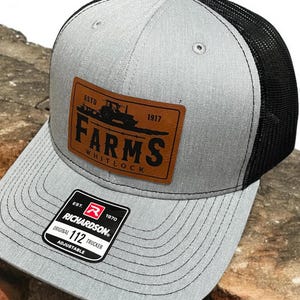 May include: A gray and black trucker hat with a brown leather patch that says "ESTD 1917 FARMS WHITLOCK". The hat is adjustable and has a Richardson 112 label.