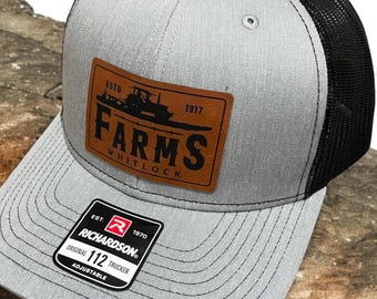 May include: A gray and black trucker hat with a brown leather patch that says "ESTD 1917 FARMS WHITLOCK". The hat is adjustable and has a Richardson 112 label.