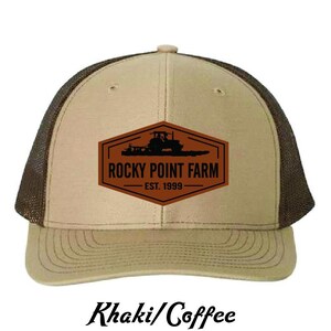 May include: Khaki and coffee-colored trucker hat with a brown mesh back. The front features a brown patch with the words "Rocky Point Farm" and a tractor graphic. The hat is labeled "Khaki/Coffee" at the bottom.