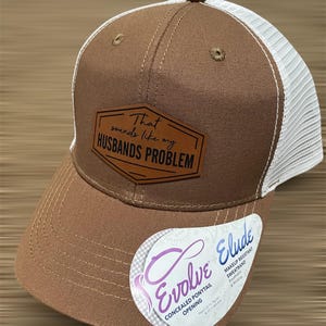 May include: Brown and white trucker hat with the text "That sounds like my Husbands Problem" on a leather patch. Features a concealed ponytail opening and makeup-resistant sweatband.