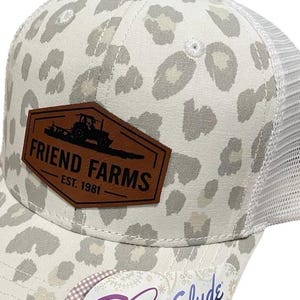 May include: A white and gray leopard print baseball cap with a mesh back. The cap features a brown leather patch with the text "FRIEND FARMS EST. 1981" and a concealed ponytail opening.