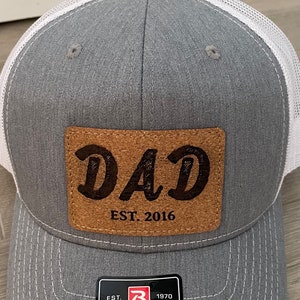 May include: Gray and white trucker hat with a brown leather patch that says "DAD EST. 2016". The hat also has a black and white label that says "EST. 1970 RICHARDSON ORIGINAL 112 TRUCKER".