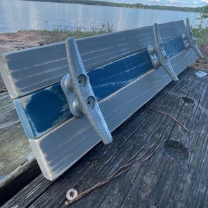 May include: A wooden coat rack with three metal hooks shaped like boat cleats. The wood is painted in shades of gray and blue.