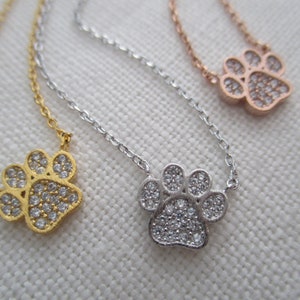 May include: Three delicate necklaces with paw print pendants. One is gold-toned, one is silver-toned, and the third is rose gold-toned. Each pendant is encrusted with small, clear crystals, and hangs from a fine chain.