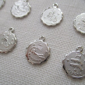 May include: A collection of silver-colored zodiac charms. Each charm features a different zodiac symbol, such as scales, a scorpion, and a pair of fish, with a textured background and a scalloped edge. Each charm has a small loop at the top.